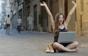 Devojka sa podignutim rukama sedi na podu sa laptopom u krilima