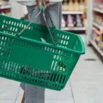 Osoba stoji ispred rafa držeći zelenu korpu.