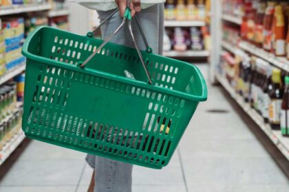 Osoba stoji ispred rafa držeći zelenu korpu.