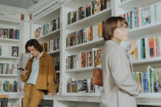 Muškarac naslonjen na policu u knjižari gleda u mobilni telefon