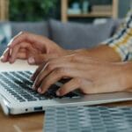Ruke kucaju na tastaturi laptopa, osoba nosi kariranu košulju