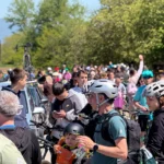 Crowd of cyclists and spectators gathered outdoors on a sunny day, many wearing helmets with bicycles in the background.