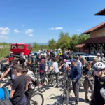 Large group of cyclists with helmets gather on a sunny road near a brick building for a biking event; some cars and a red truck in the background.