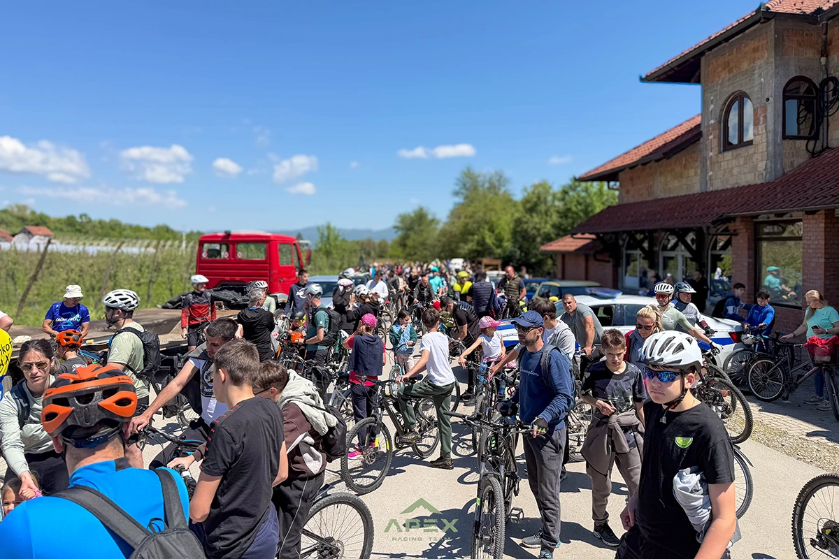 Large group of cyclists with helmets gather on a sunny road near a brick building for a biking event; some cars and a red truck in the background.