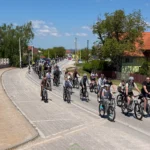 Group of cyclists riding on a sunny rural road past houses and greenery.