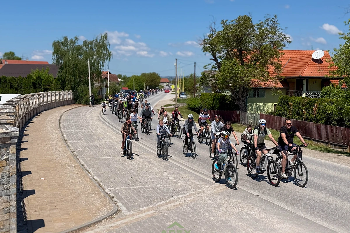 Group of cyclists riding on a sunny rural road past houses and greenery.