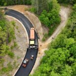 Aerial view of road construction in a forest: curved black asphalt with orange trucks laying pavement.