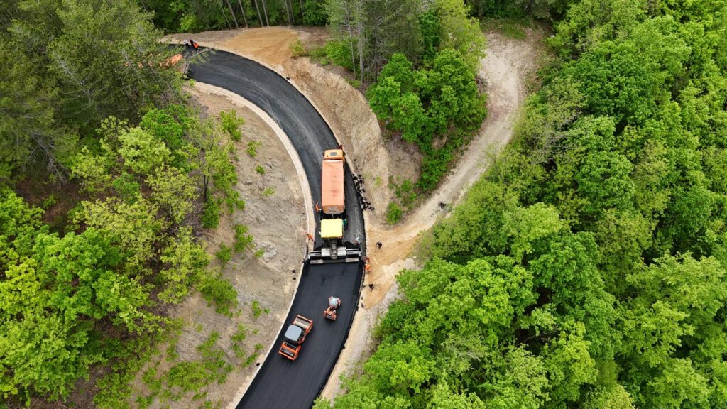 Aerial view of road construction in a forest: curved black asphalt with orange trucks laying pavement.