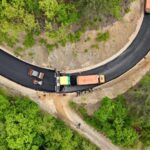 Aerial view of a curved, freshly paved road under construction in a forest, with trucks and workers along the center.