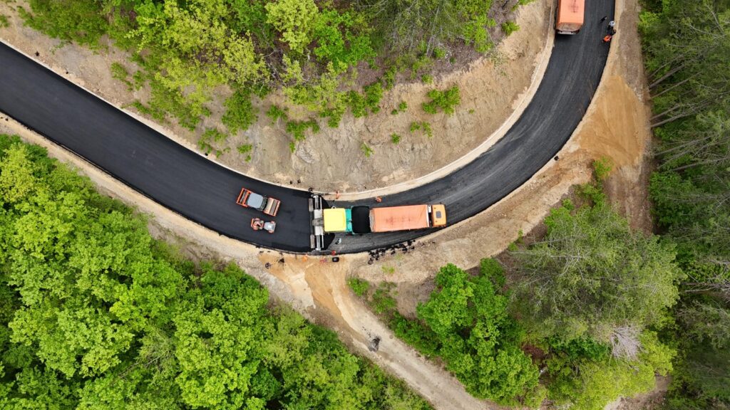 Aerial view of a curved, freshly paved road under construction in a forest, with trucks and workers along the center.