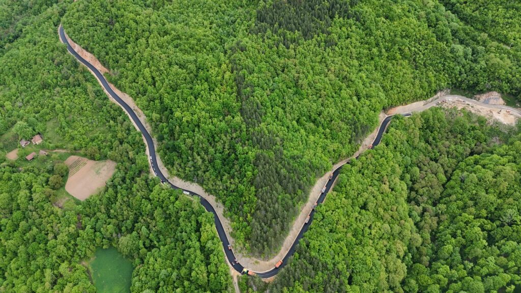 Aerial view of a long, winding dark road or barrier cutting through dense green forest with small clearings nearby.