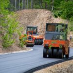 Orange road rollers compacting fresh asphalt on a winding forest road surrounded by rocky slopes and green trees.