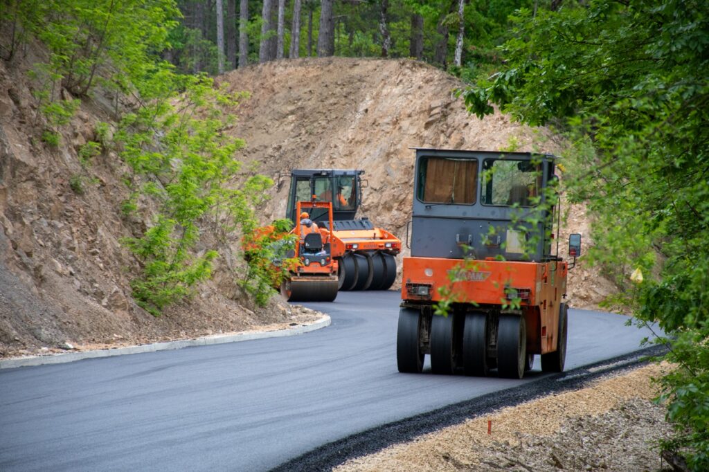 Orange road rollers compacting fresh asphalt on a winding forest road surrounded by rocky slopes and green trees.