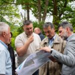 Group of men standing outdoors in a forested area, examining a large map or brochure together.