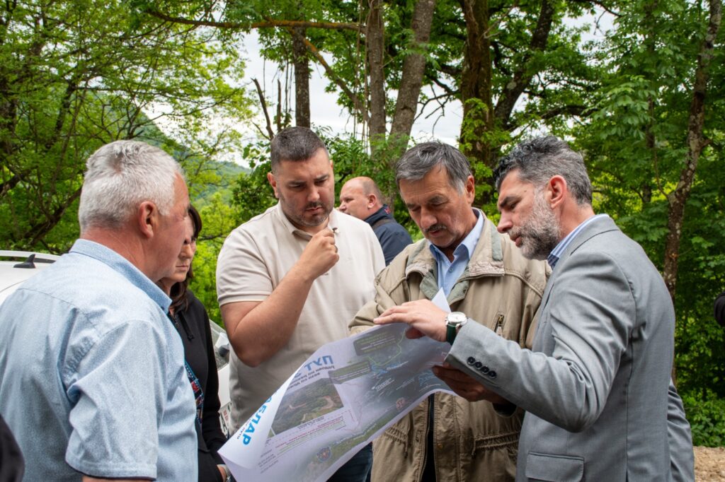 Group of men standing outdoors in a forested area, examining a large map or brochure together.