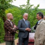 Three men stand in a green outdoor setting near a red car, talking together; one in a plaid jacket, one in a gray suit, and another in a beige jacket.