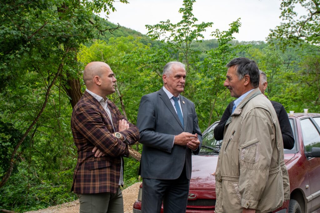Three men stand in a green outdoor setting near a red car, talking together; one in a plaid jacket, one in a gray suit, and another in a beige jacket.