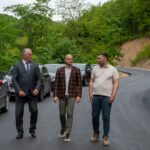 Three men walk along a freshly paved road in a forested area, with parked cars on the left.