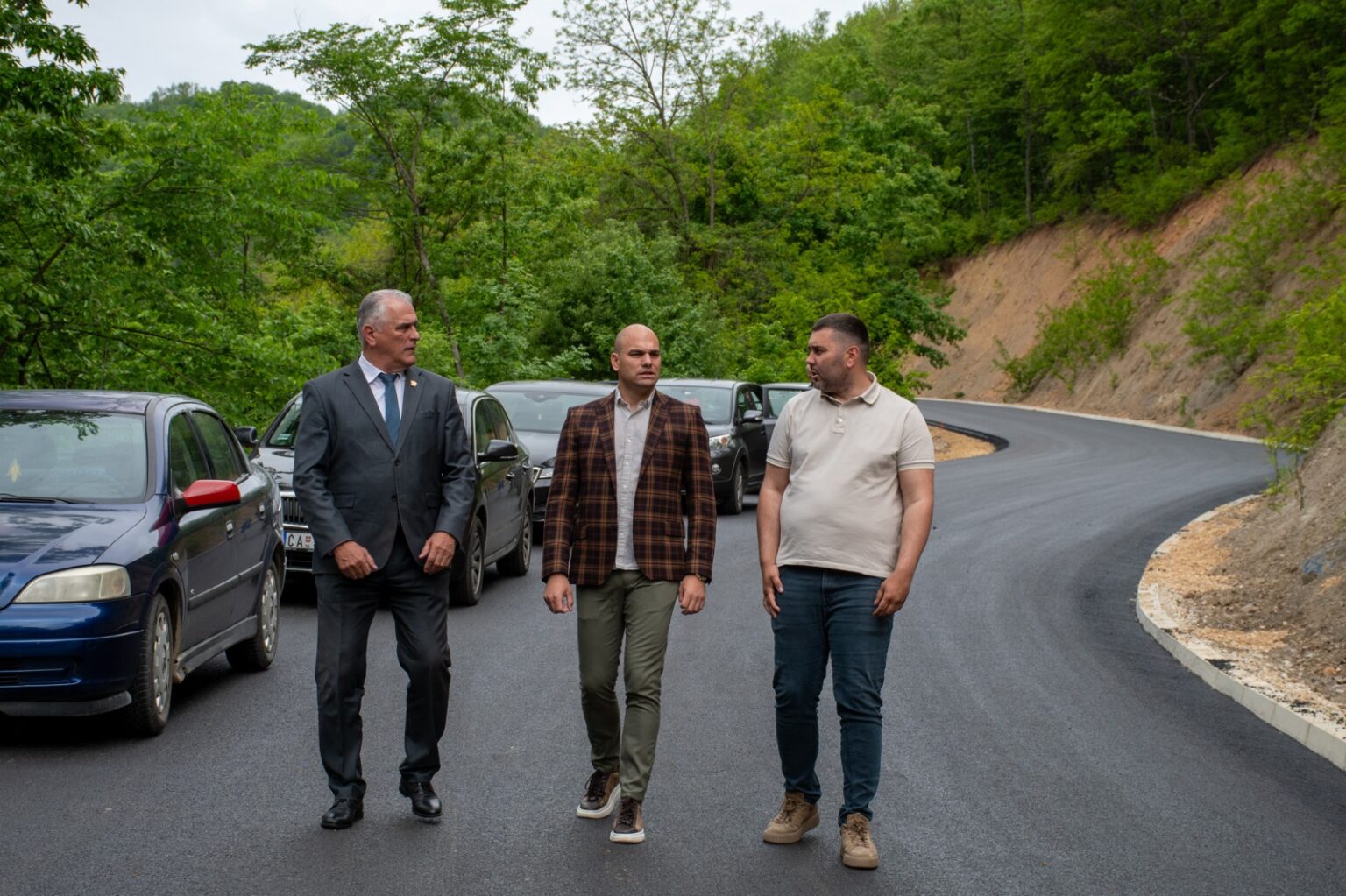 Three men walk along a freshly paved road in a forested area, with parked cars on the left.