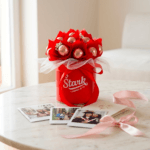Red bouquet in a red tin with white tulle, placed on a white marble table; photos and pink ribbon nearby suggest a gift scene.