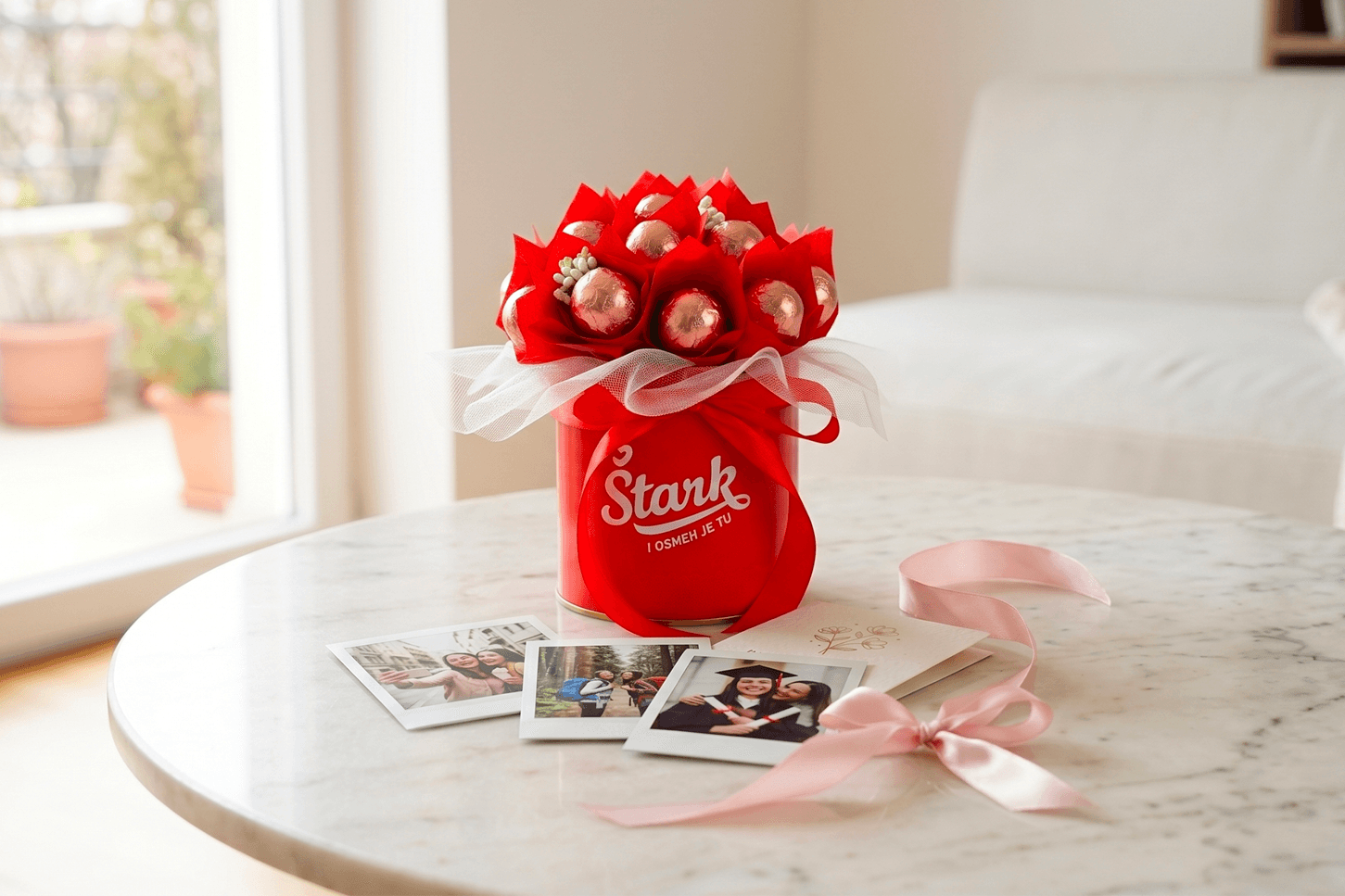 Red bouquet in a red tin with white tulle, placed on a white marble table; photos and pink ribbon nearby suggest a gift scene.