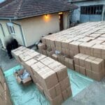 Outdoor area with many stacked brown cardboard boxes on a tarp, near a beige building; blurred figures nearby in the left foreground.