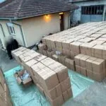 Outdoor area with many stacked brown cardboard boxes on a tarp, near a beige building; blurred figures nearby in the left foreground.