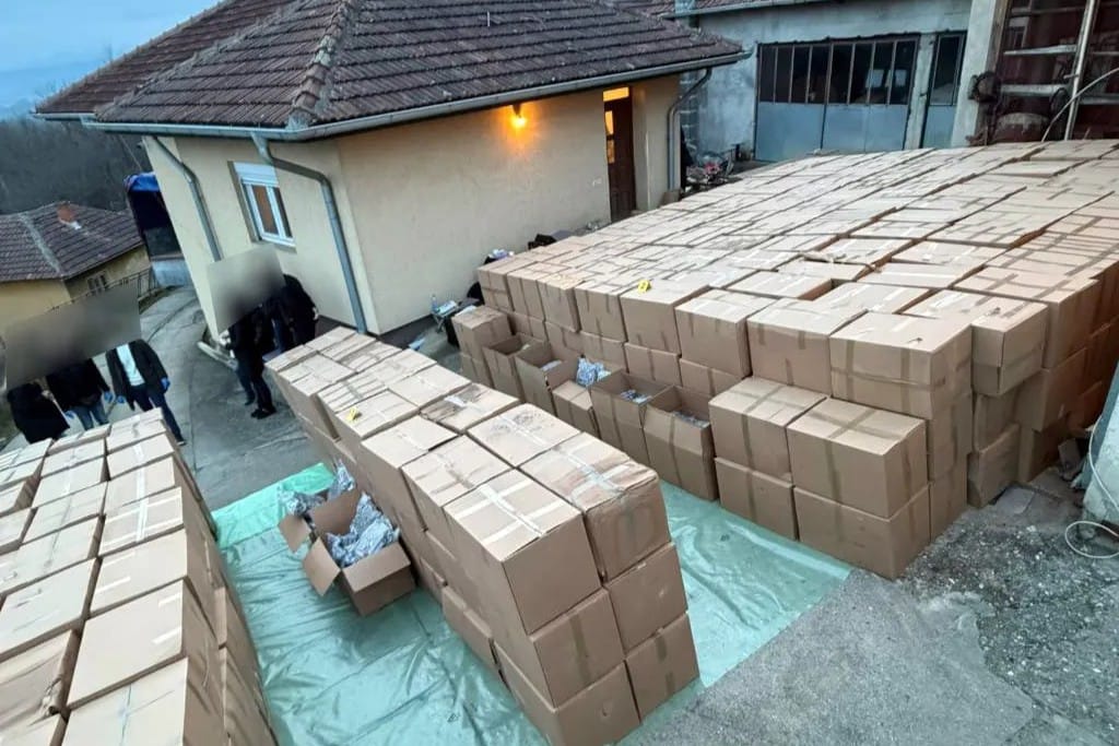 • N2 portal Outdoor area with many stacked brown cardboard boxes on a tarp, near a beige building; blurred figures nearby in the left foreground.